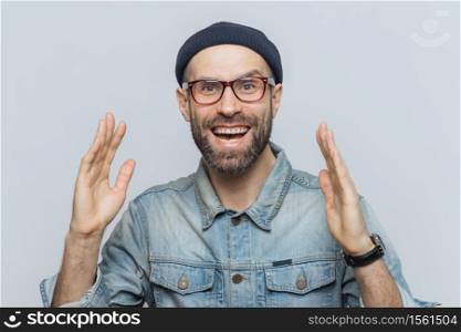 Handsome happy man raises hands with excitment, has overjoyed expression, looks with delighted face, has thick beard and mustache. Glad man in eyewear poses against white background, has fun