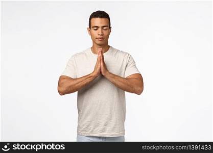 Handsome healthy muscular male in t-shirt, clasp hands together over chest, close eyes and meditate, practice yoga, praying with faithful happy expression, inhale fresh air, trying find peace in mind.. Handsome healthy muscular male in t-shirt, clasp hands together over chest, close eyes and meditate, practice yoga, praying with faithful happy expression, inhale fresh air, trying find peace in mind