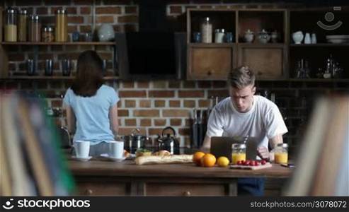 Handsome hipster browsing social media on laptop while his girlfriend preparing breakfast in the kitchen. Young couple spending leisure together in the morning at home.