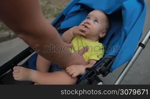 Handsome hipster dad with beard picking up his infant child from baby stroller and kissing while spending time together in public park. Affectionate father bonding with toddler baby boy over beautiful landscape background. Slo mo. Stabilized shot.
