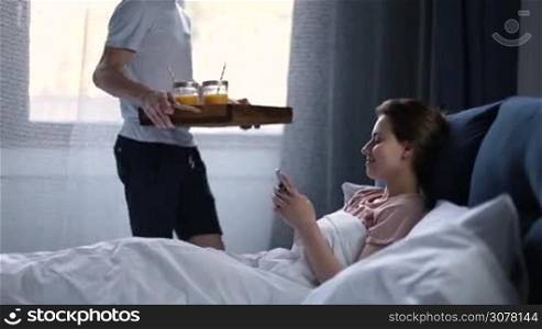 Handsome hipster surprising his beautiful and happy girlfriend with breakfast in bed, bringing juice in mason jars and croissants in wooden tray. Young man serving romantic breakfast to his woman in bed in the morning.