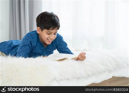 Handsome Indian boy in traditional clothing lying on bed smiling and reading book, having fun on free time at home. Education concept
