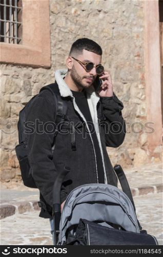 Handsome looking man with golden framed sunglasses behind a baby stroller talks on his phone in a sunny cobblestone street. Travel concept.. Man behind a baby stroller talks on his phone