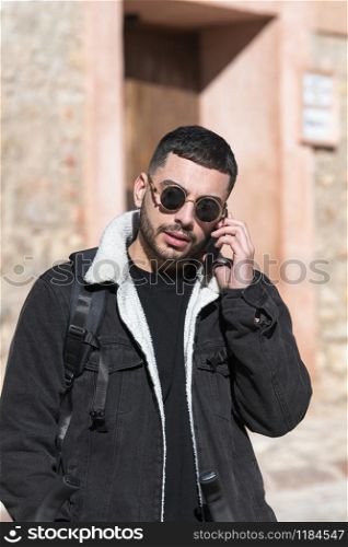 Handsome looking with golden framed sunglasses talks on his phone with a hand in his pocket on an out of focus background. Travel concept.. Handsome man talks on his phone in a rustic street
