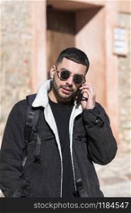 Handsome looking with golden framed sunglasses talks on his phone with a hand in his pocket on an out of focus background. Travel concept.. Handsome man talks on his phone in a rustic street