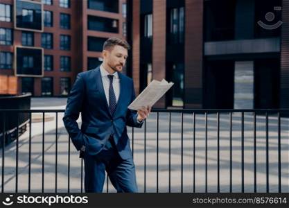 Handsome male boss wearing blue suit, reading newspaper thoughtfully, looking for news regarding his firm, standing alone with one hand in his pocket in front of corporate buildings. Boss reading newspaper in front of corporate building