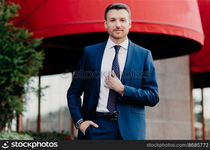 Handsome male company owner dressed formally, stands outside, has confident expression, ready for business meeting. Successful young attractive executive manager poses outdoor near office building