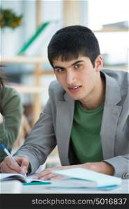 Handsome male student looking at camera while making notes sitting at classroom with other students