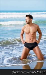 Handsome male with muscular naked torso and in swimming trunks standing on knees in sea water on beach and looking away. Fit man in shorts in sea water
