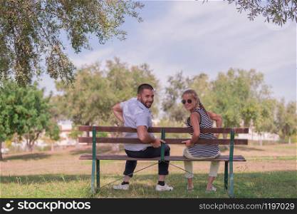 Handsome man and beautiful woman sitting in a bench face backwards and smile while looking at the camera on an out of focus nature background. Leisure concept.. Smiling man and woman sitting on a bench