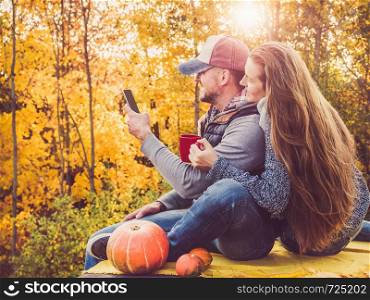 Handsome man and stylish woman holding a mobile phone and sitting on the terrace against the background of yellow trees and the setting sun. Happy relationship concept. Handsome man and stylish woman holding a phone