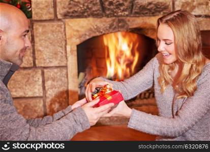Handsome man giving gift to his beautiful wife, spending Christmas holidays in cozy house near fireplace, love and romance concept
