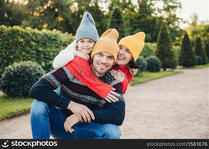 Handsome man has good relationship with wife and small daughter who embrace him from back, have pleasant smiles. Friendly young family in knitted hats and sweaters stand against green trees