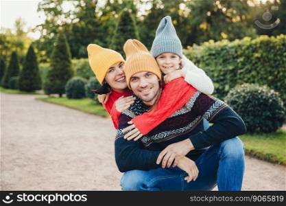 Handsome man has good relationship with wife and small daughter who embrace him from back, have pleasant smiles. Friendly young family in knitted hats and sweaters stand against green trees