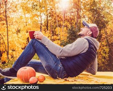 Handsome man holding a red mug and sitting on the terrace against the background of yellow trees and the setting sun. Happy relationship concept. Handsome man holding a red, stylish mug