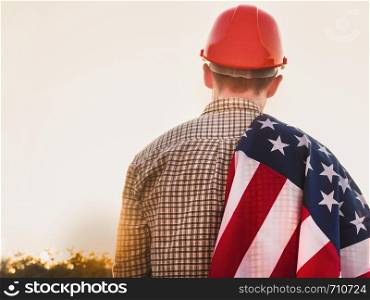 Handsome man holding an American Flag, standing against a background of green trees and the rays of the setting sun. View from the back, close-up. Concept of work and employment. Beautiful card with congratulations on Labor Day