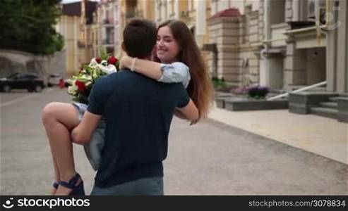 Handsome man holding cute smiling girlfriend in his arms and spinning around on street. Cheerful attractive woman with bouquet of awesome flowers in loving embrace of her boyfriend Slow motion.