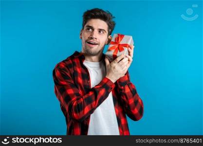 Handsome man holding gift box on blue studio background smiles to camera. Happy european guy received present and interested in what&rsquo;s inside. Handsome man holding gift box on blue studio background smiles to camera. 