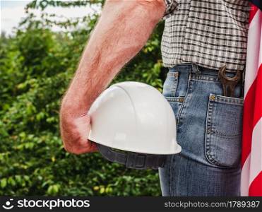 Handsome man holding US Flag and construction helmet against the background of trees, blue sky and sunset. View from the back. Labor and employment concept. Attractive man holding construction helmet in his hands