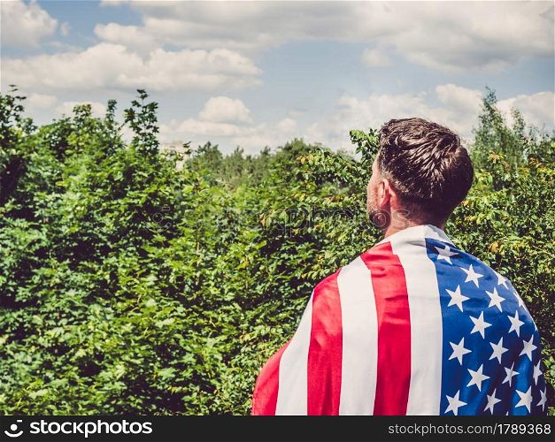 Handsome man holding US flag in his hands against the background of trees, blue sky and sunset. View from the back. Labor and employment concept. Handsome man holding US flag in his hands