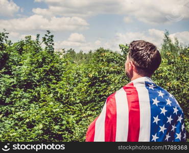 Handsome man holding US flag in his hands against the background of trees, blue sky and sunset. View from the back. Labor and employment concept. Handsome man holding US flag in his hands