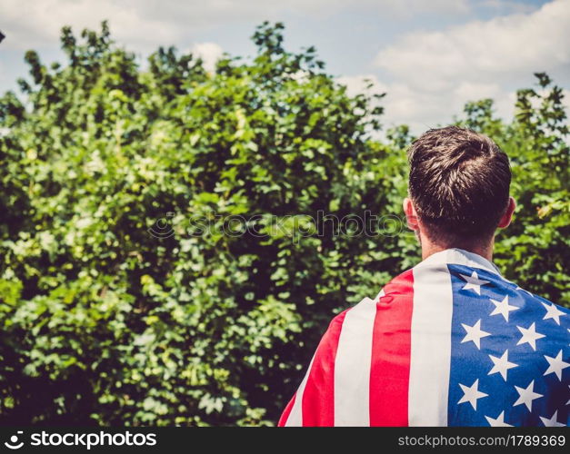 Handsome man holding US flag in his hands against the background of trees, blue sky and sunset. View from the back. Labor and employment concept. Handsome man holding US flag in his hands