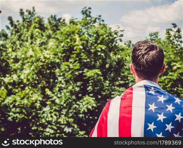 Handsome man holding US flag in his hands against the background of trees, blue sky and sunset. View from the back. Labor and employment concept. Handsome man holding US flag in his hands
