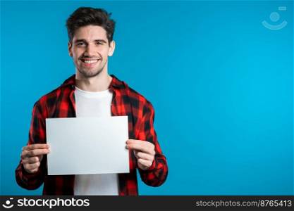Handsome man holding white a4 paper poster. Copy space. Smiling hipster guy in red plaid shirt on blue background. Handsome man holding white a4 paper poster. Copy space. Smiling hipster guy in red plaid shirt on blue background.