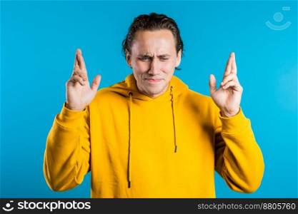 Handsome man in yellow hoodie praying, crossing fingers over blue background. European race guy begging someone. Religion concept. High quality photo. Handsome man in yellow hoodie praying, crossing fingers over blue background. European race guy begging someone. Religion concept.