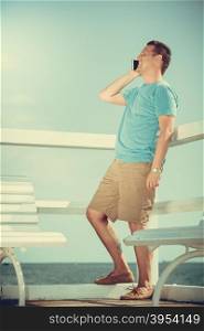 Handsome man tourist on pier talking on phone. Fashion guy enjoying summer travel vacation by sea. Relax and technology concept.. Man tourist on pier using smartphone. Technology.
