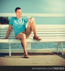 Handsome man tourist sitting on bench on pier. Guy enjoying summer travel vacation by sea. Fashion and relaxation.. Handsome man tourist on pier. Fashion summer.