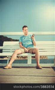 Handsome man tourist sitting on bench on pier using smartphone. Fashion guy enjoying summer travel vacation by sea. Relax and technology concept.. Man tourist on pier using smartphone. Technology.