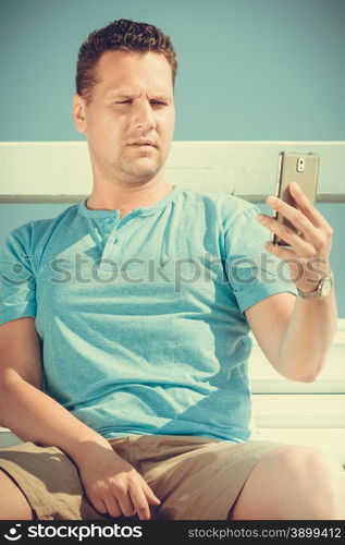 Handsome man tourist sitting on bench on pier using smartphone. Fashion guy enjoying summer travel vacation by sea. Relax and technology concept.. Man tourist on pier using smartphone. Technology.