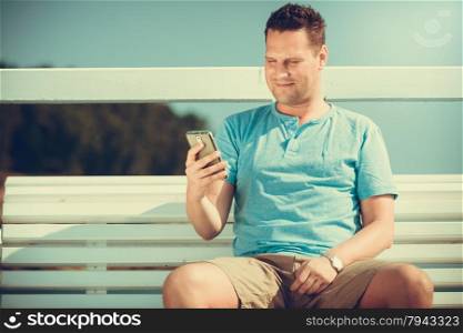 Handsome man tourist sitting on bench on pier using smartphone. Fashion guy enjoying summer travel vacation by sea. Relax and technology concept.. Man tourist on pier using smartphone. Technology.