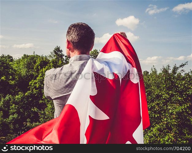 Handsome man waving a Canadian Flag against a background of trees and blue sky. View from the back, close-up. National holiday concept. Man waving a Canadian Flag. National holiday