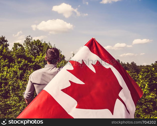 Handsome man waving a Canadian Flag against a background of trees and blue sky. View from the back, close-up. National holiday concept. Man waving a Canadian Flag. National holiday