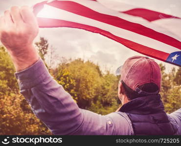 Handsome man waving an American flag against a background of trees and blue sky. View from the back, close-up. National holiday concept. Handsome, young man waving an American flag