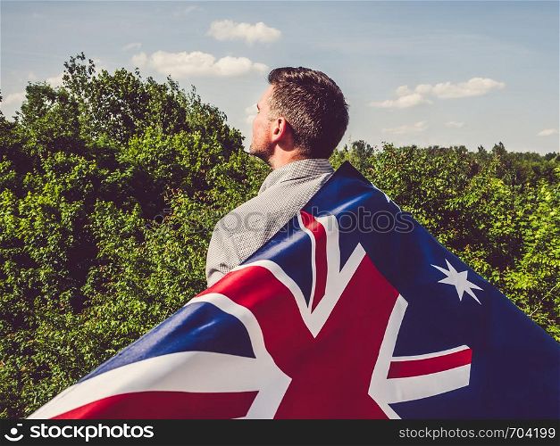 Handsome man waving the Flag of Australia against the backdrop of trees and blue sky. View from the back, close-up. National holiday concept. Handsome man waving the Flag of Australia