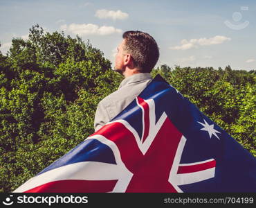 Handsome man waving the Flag of Australia against the backdrop of trees and blue sky. View from the back, close-up. National holiday concept. Handsome man waving the Flag of Australia