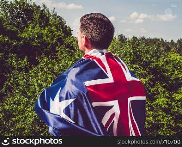 Handsome man waving the Flag of Australia against the backdrop of trees and blue sky. View from the back, close-up. National holiday concept. Attractive, young man waving a British Flag