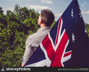 Handsome man waving the Flag of Australia against the backdrop of trees and blue sky. View from the back, close-up. National holiday concept. Attractive, young man waving a British Flag