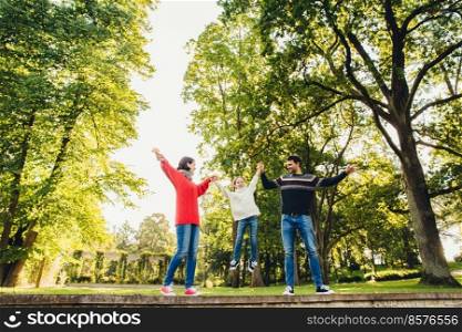 Handsome man wears sweater and jeans, beautiful woman in red knitted sweater and little adorable girl keeps with hands her parents, stand against beautiful nature background. Spending time together