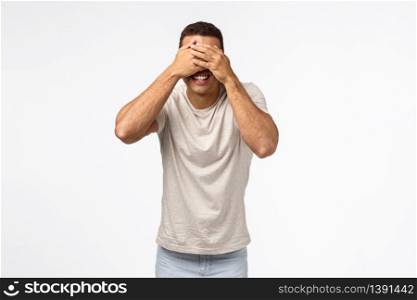 Handsome masculine young man playing hide-n-seek or peekaboo, cover eyes, standing blindfolded and smiling happy, waiting for signal to look at surprise christmas gift, white background.. Handsome masculine young man playing hide-n-seek or peekaboo, cover eyes, standing blindfolded and smiling happy, waiting for signal to look at surprise christmas gift, white background