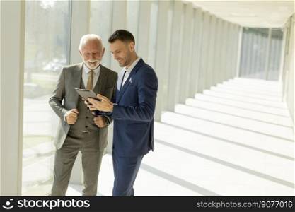 Handsome mature businessman and his young colleague discussing finantial report on   digital tablet in the office corridor