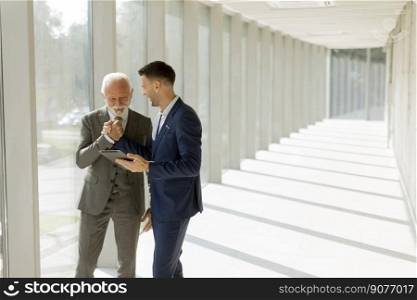 Handsome mature businessman and his young colleague discussing finantial report on   digital tablet in the office corridor