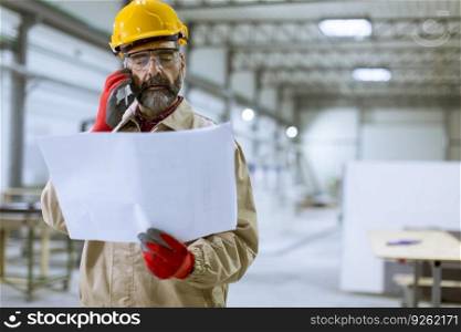 Handsome mature engineer looking at plan and using mobile phone in the factory