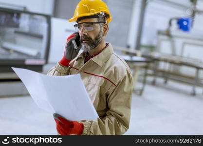 Handsome mature engineer looking at plan and using mobile phone in the factory