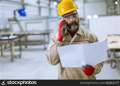 Handsome mature engineer looking at plan and using mobile phone in the factory