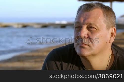 Handsome middle-aged man sitting thinking in the sunlight at the coast with a serious expression and ocean backdrop