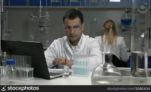 Handsome mixed race male scientific researcher examining test tubes with liquid and recording results of experiment on latop in chemical lab. Male scientist in protective goggles working in laboratory with coworker on background.